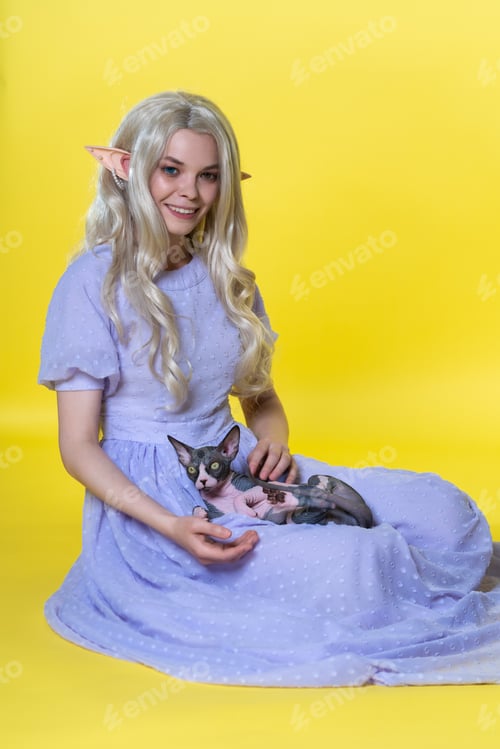 Preview: Cosplayer elf young blonde woman in blue dress sits on yellow background, holds Sphinx cat on lap