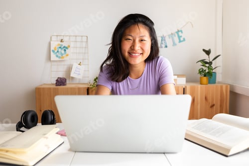 Preview: Happy teen asian female college student studying at home using laptop looking at camera.