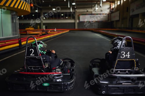 Preview: two diverse racers in helmets and sportswear driving go kart on indoor circuit, competitors