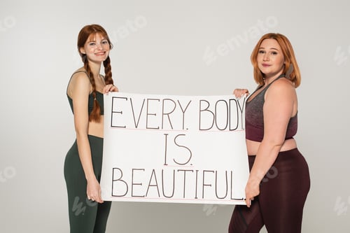 Preview: Smiling sportswoman with freckles holding placard with every body is beautiful lettering near plus
