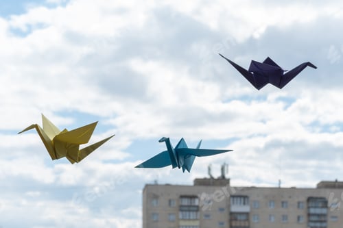 Preview: Multicolored paper origami bird cranes hanging on the window against sky. Origami crane