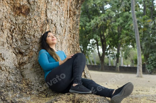 Preview: Woman seated leaning against a tree with pensive attitude.