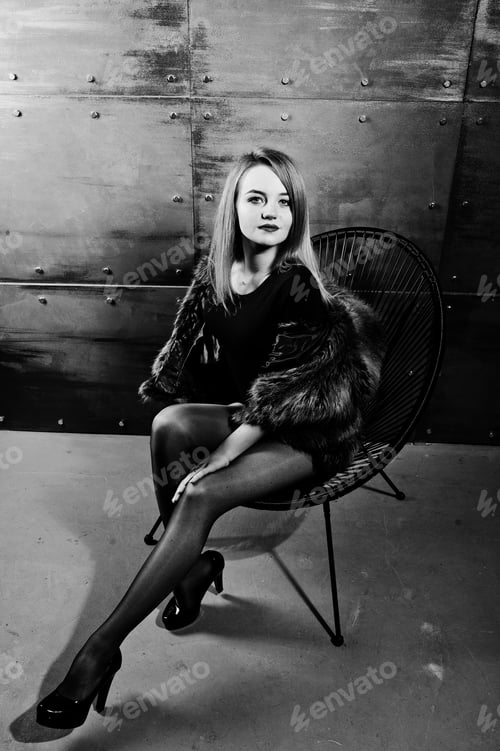 Visualização: Menina loira elegante usa casaco de pele e vestido combinado posado na cadeira contra a parede de aço no estúdio.