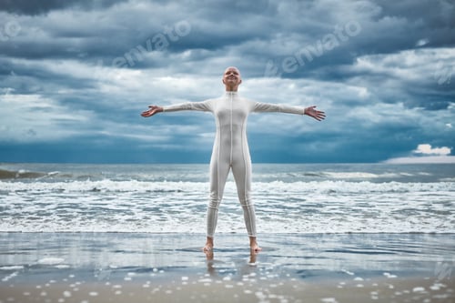 Preview: Happy hairless girl with alopecia in white futuristic suit stands on beach bathed by ocean waves