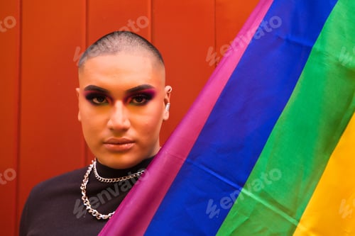 Preview: Hispanic boy with vibrant makeup holding gay pride flag against red wooden door