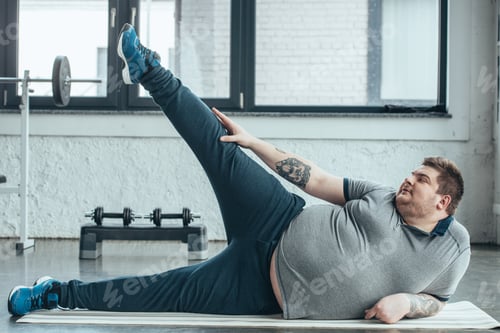 Preview: overweight tattooed man lying on fitness mat and stretching legs at gym