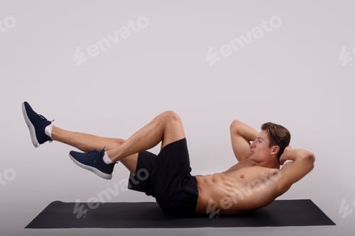 Preview: Strong young guy with bare torso doing abs exercises on yoga mat against light studio background