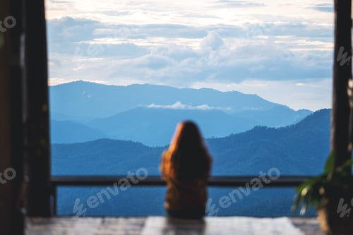 Preview: Blurred rear view image of a female traveler sitting and looking at a beautiful mountain