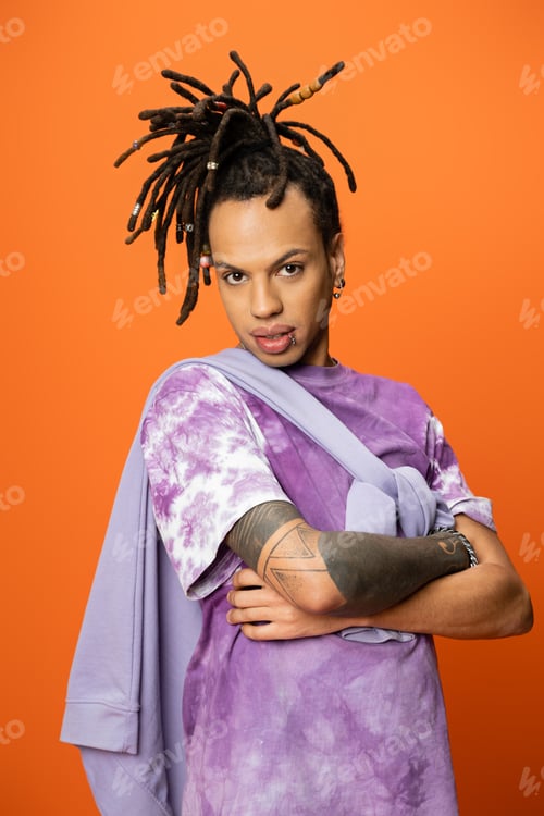 Preview: trendy multiracial man with dreadlocks posing with crossed arms isolated on orange
