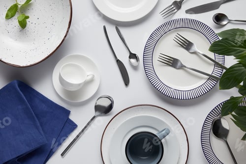 Preview: top view of green leaves, different ceramic plates, cup, kitchen towel, forks, spoons and knives on