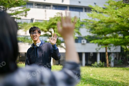 Preview: Happy Asian male college student waves his hand to greet his friend at the campus park.