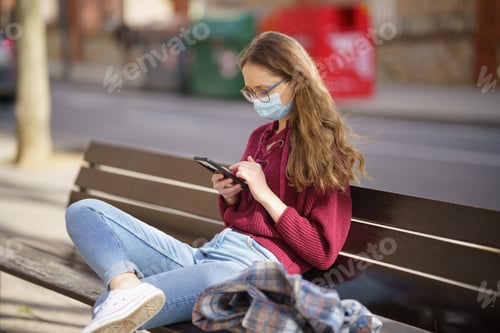 Preview: Young Caucasian woman in a mask sitting on the bench and looking at her phone