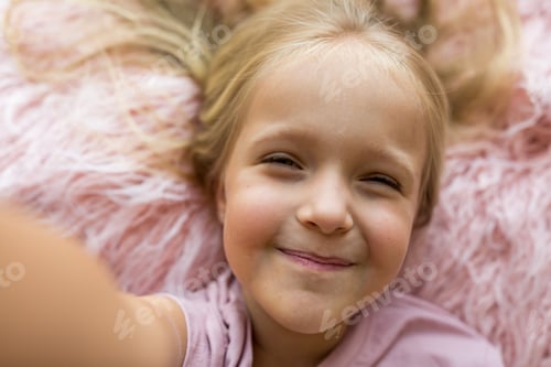 Preview: Happy Caucasian little girl 5-6 years old with blonde hair making selfie at home on bed