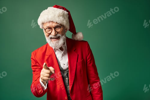 Preview: joyful elegant Santa with beard smiling cheerfully and pointing finger at camera, winter concept