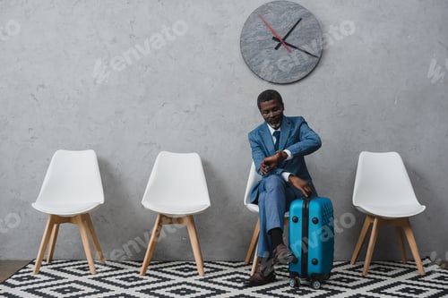 Preview: Handsome African american businessman sitting in a waiting room with suitcase and looking at watch
