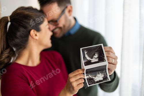 Preview: Happy young couple, pregnant woman, holding and showing ultrasound scan picture at home