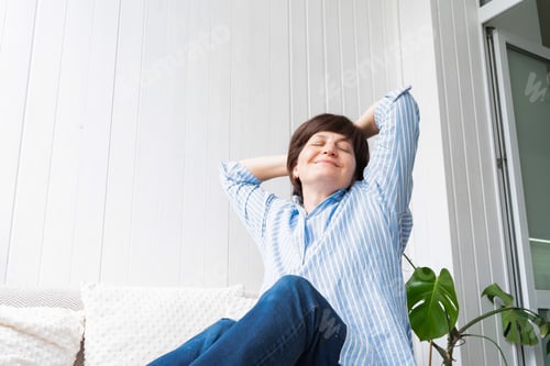 Preview: Relaxed adult woman enjoying rest in bright room sitting on light sofa with light soft pillows