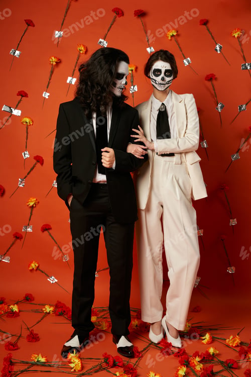 Preview: full length of couple in catrina makeup and festive attire posing on red backdrop with carnations