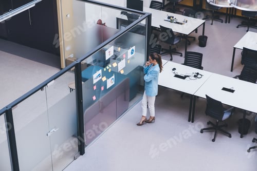 Preview: Young focused woman standing by glass wall with sticky notes in office