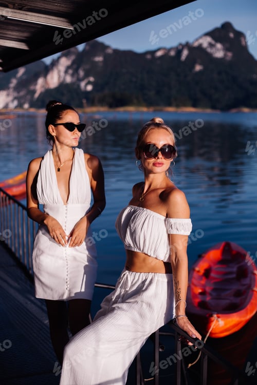 Preview: Two stylish woman in white summer clothes wearing sunglasses lake on background
