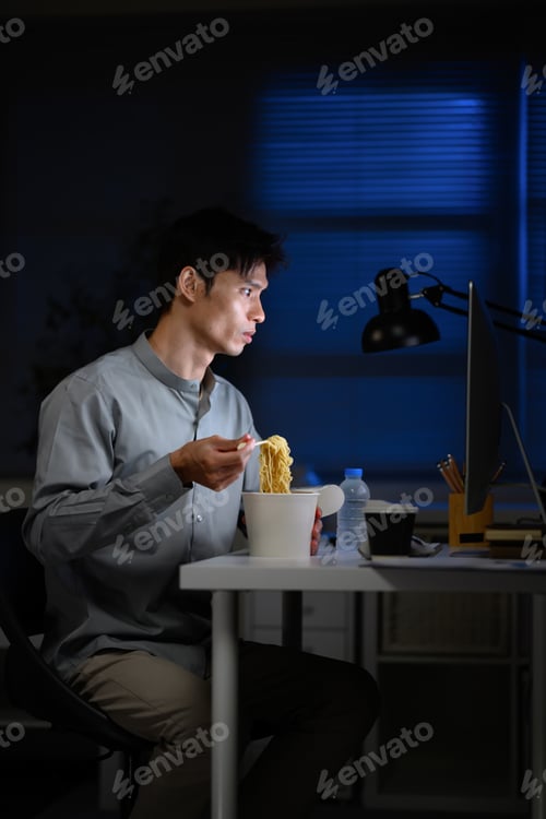 Preview: Asian businessman eating instant noodles while working late at office