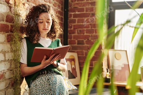 Preview: Young serious woman in casualwear holding open book and reading it by brick wall