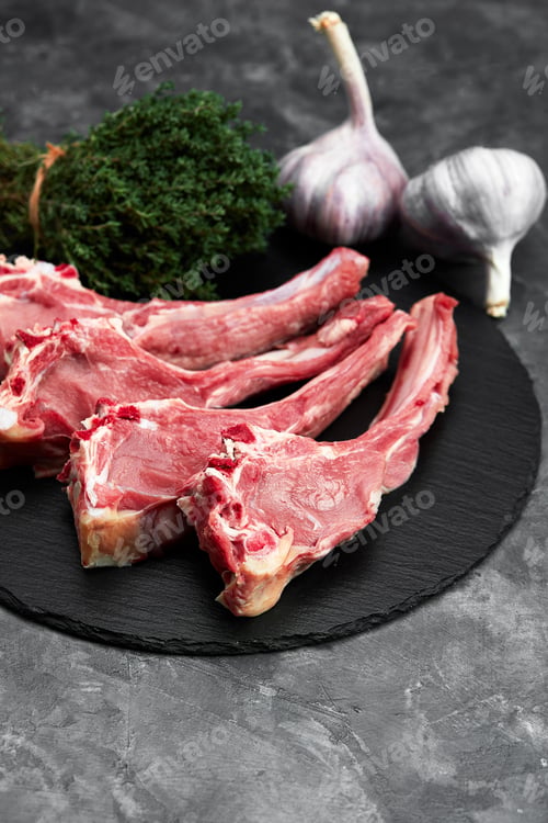 Preview: Raw lamb chops with garlic, herbs on black ceramic plate over white stone gray table. Top view. With