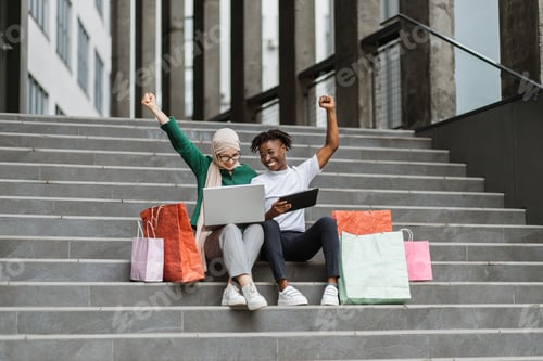 Preview: Multiethnical girls sitting on steps of a modern building rejoicing with online orderafter shopping