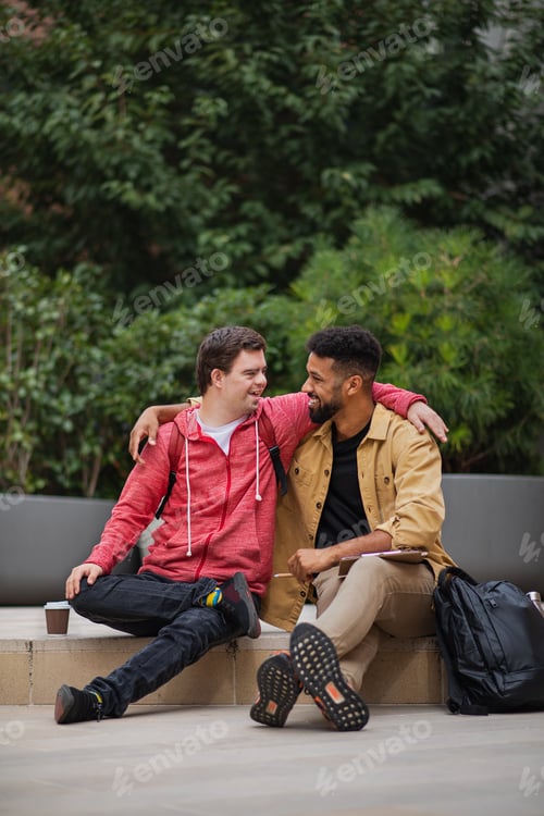 Preview: Happy young man with Down syndrome and mentoring friend sitting with arms around outdoors