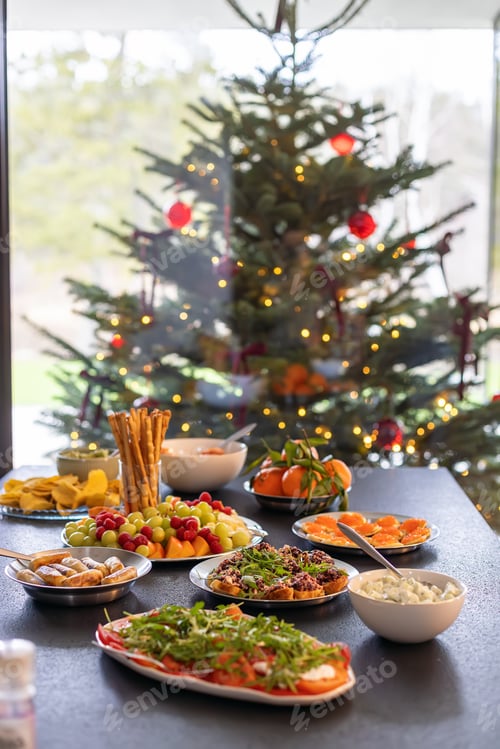 Preview: A black table fully set with appetizers, fruit, crackers, and dips, in front of a Christmas tree