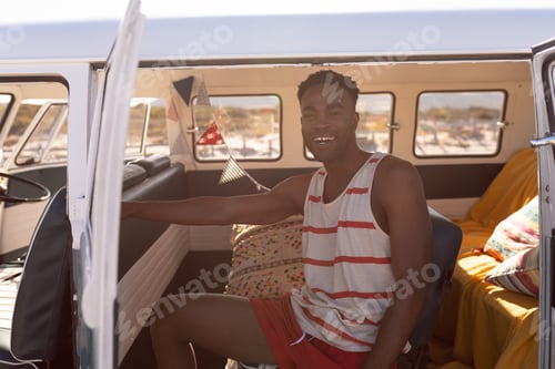Preview: front view of happy young African-american man sitting in camper van at beach