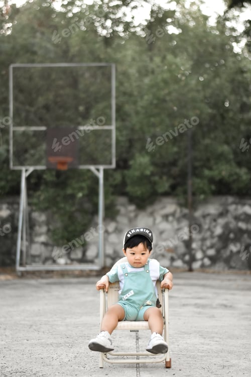 Preview: Little boy in a cap and pants with suspenders sits on an chair in park