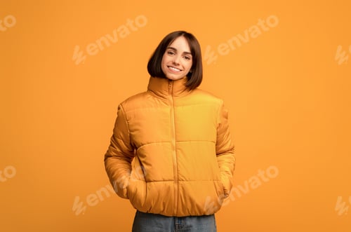 Preview: Winter clothing. Young smiling woman in warm jacket, holding hands in pockets and looking at camera