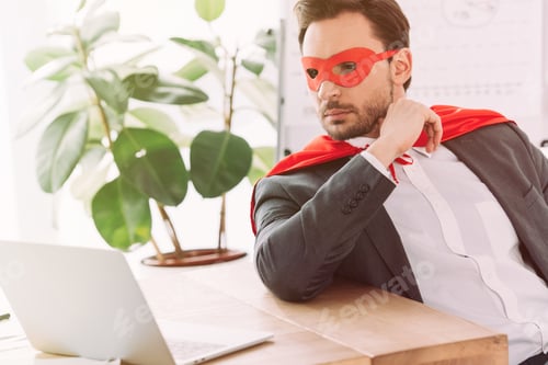 Preview: handsome super businessman in mask and cape looking at laptop in office