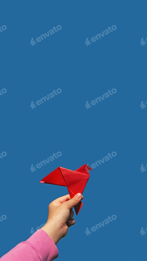 Preview: red paper origami pigeon in children's hand on blue sky background, vertical, 16:9, copy space