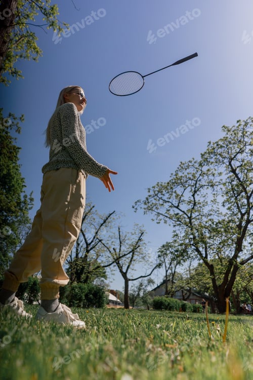 Preview: outdoors in a park with trees young blonde girl stands with a bambinton racket posing for the camera