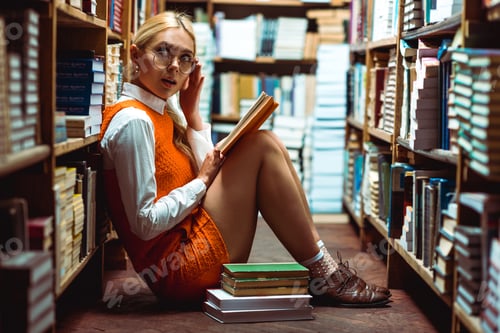 Preview: beautiful and blonde woman in glasses holding book and sitting on floor in library
