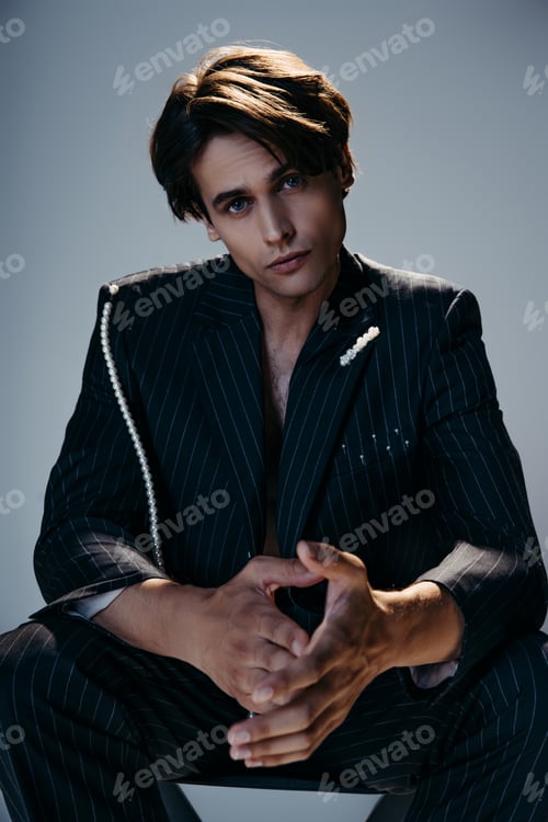 Preview: stylish man in trendy suit with pearl chain sitting while looking at camera on dark grey