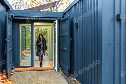 Preview: Businesswoman walking out modular shipping container office with solar panels in autumn