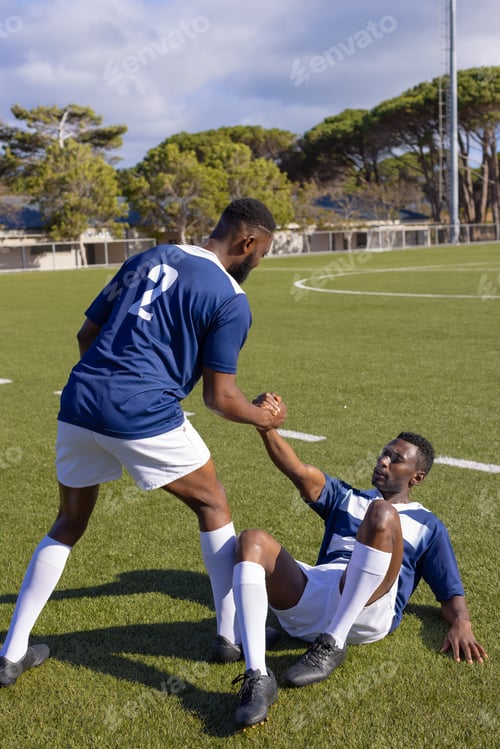 Preview: Two African American young male athletes are on a rugby field outdoors, one helping the other up