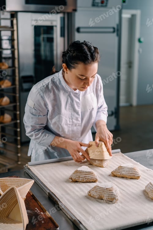 Preview: concentrated beautiful woman baker lays out raw bread rolls from molds before baking
