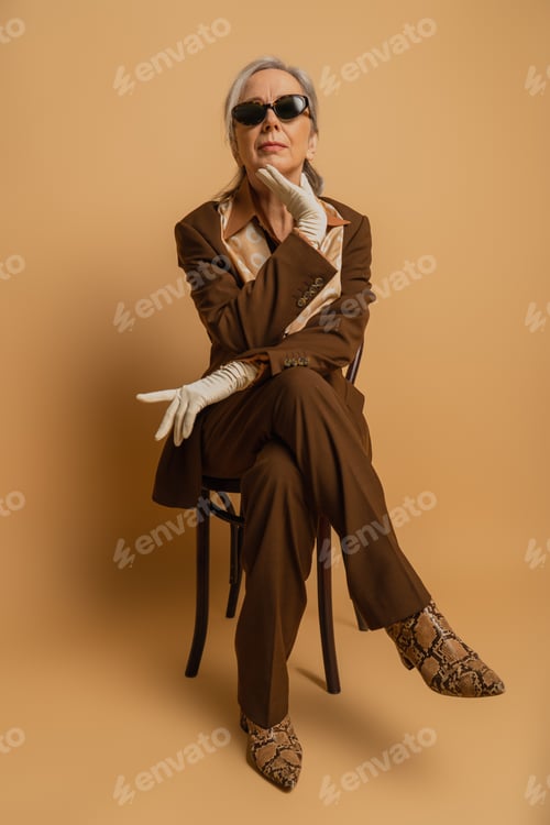 Preview: full length of senior woman in brown formal wear and sunglasses sitting on chair and looking at