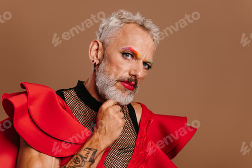Preview: Portrait of confident mature gay man in red dress looking at camera against brown background