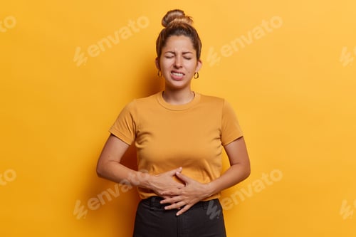 Preview: Photo of unhappy European woman with hair bun keeps hands on stomach suffers from pain or pms