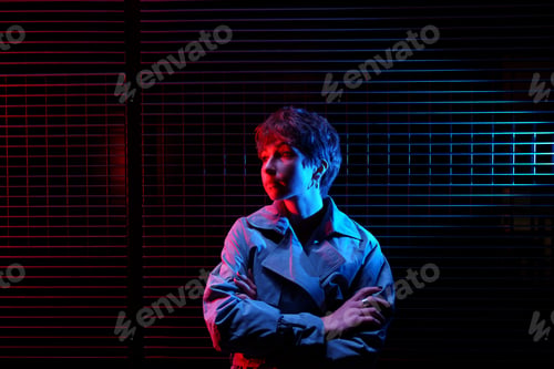 Preview: Girl illuminated by colourful light stands near wall with arms crossed and looks thoughtful to side.