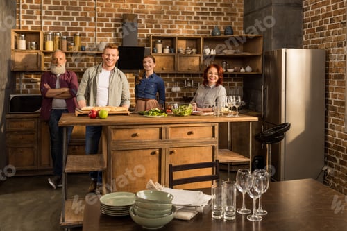 Preview: happy multigeneration family looking at camera while standing in kitchen together at home