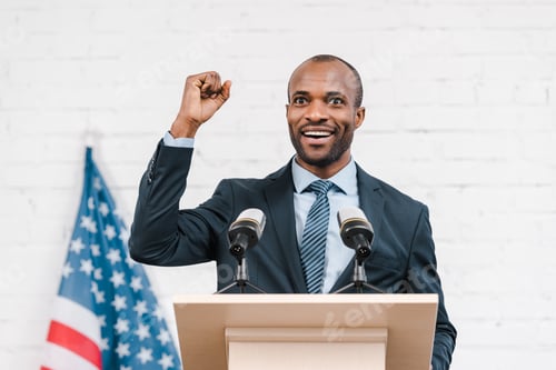 Preview: happy african american speaker with clenched fist smiling near microphones and american flag