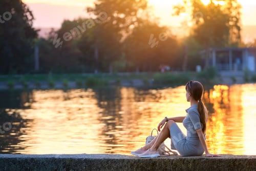 Preview: Back view of lonely young woman sitting alone on lake shore enjoying warm evening.