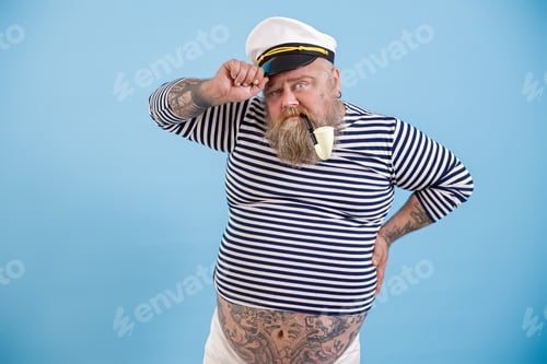 Preview: Mature fat sailor with beard and vintage pipe adjusts hat on light blue background