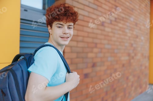 Preview: Happy teen boy with backpack walks by brick wall on a sunny morning